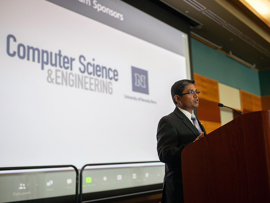 Shamik Sengupta, dressed in a suit and standing at a podium with a large screen behind him that has the logo for the College of Engineering's Computer Science & Engineering Department.
