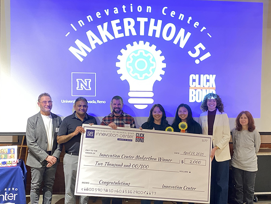 Group of seven individuals posing with a large check at the University of Nevada, Reno Innovation Center Makerthon 5 event, under a lit-up sign reading "Makerthon 5! Click Bond.