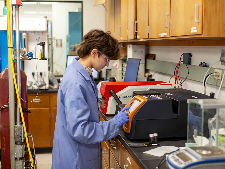 Jade Beland, wearing safety goggles and a lab coat, operates scientific equipment in Krista Carlson's lab.