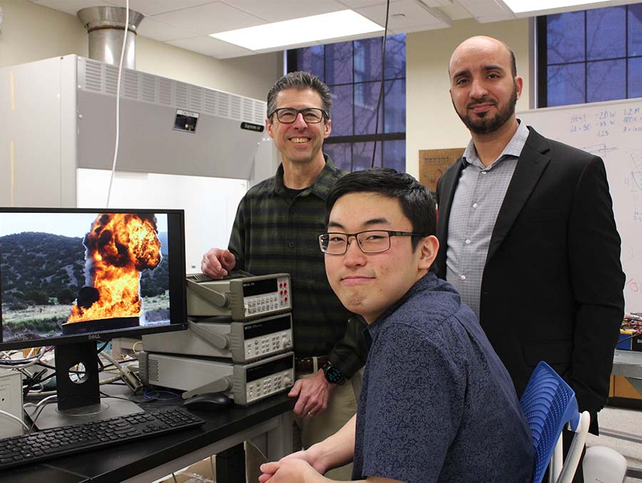 Three researchers in a laboratory stand behind a seated student working at a computer, which displays an image of an explosion outdoors.